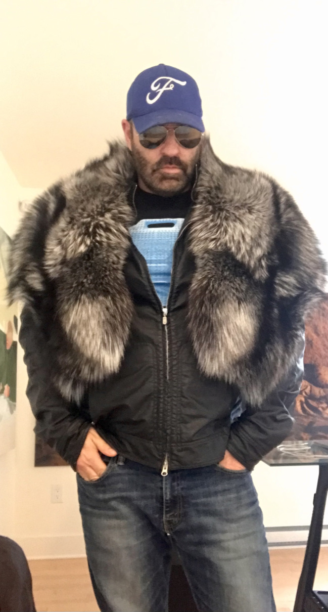 man wearing wide silver fox fur pelerine, sunglasses and cap, hands in pockets
